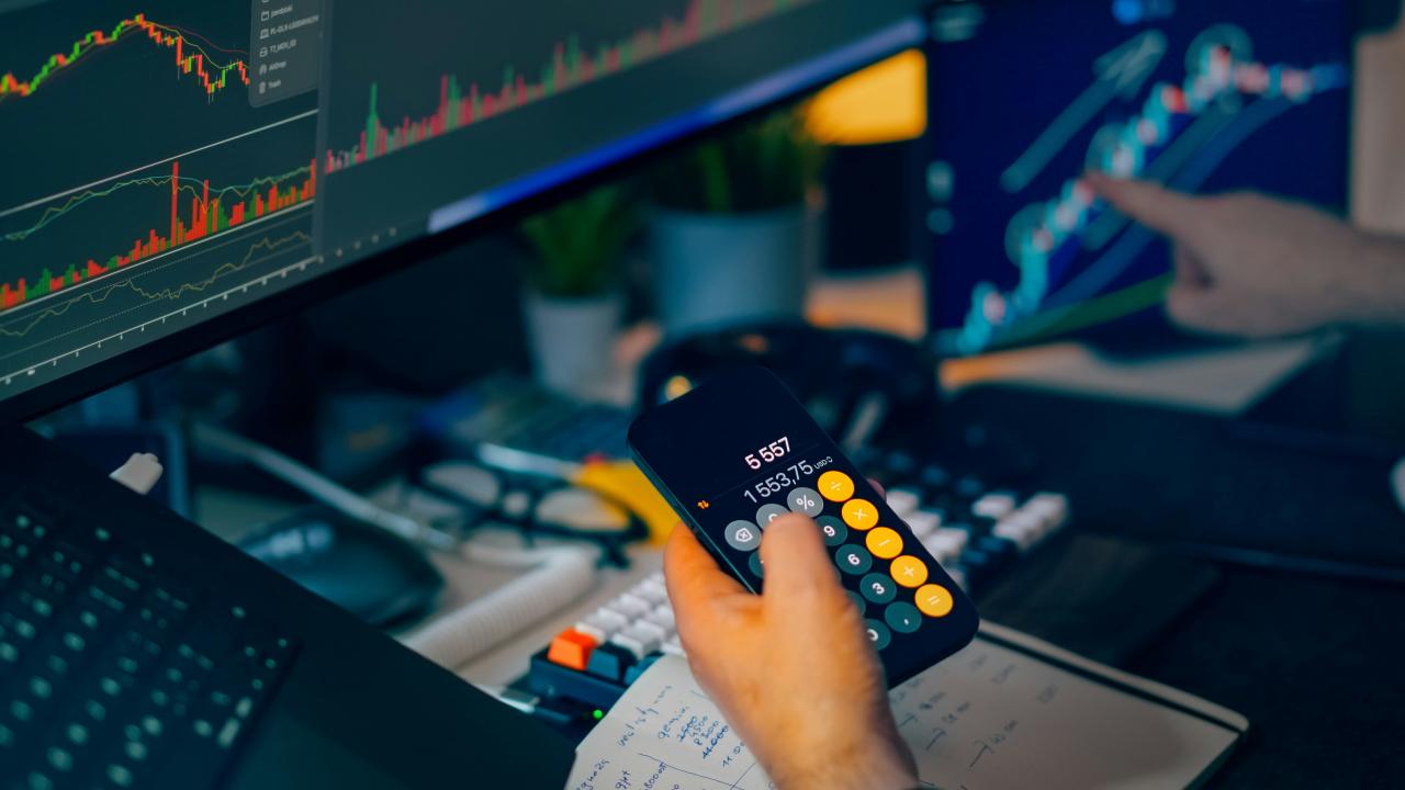 Trader analyzing stock market charts on computer screens with calculator.